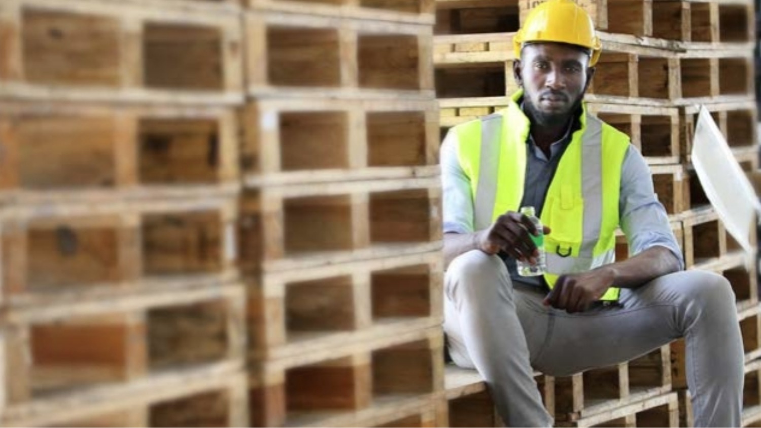 African American worker in safety vest suit and hardhat is drinking water during break while sitting on the pile of pallet wood at warehouse 